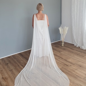 Puede incluir: Un velo de novia de tul blanco con una larga cola. El velo está drapeado sobre un vestido blanco y la modelo está de espaldas a la cámara.