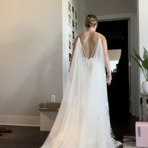 May include: A white wedding dress with a long train and a lace overlay. The dress has a low back and a cape-like overlay.