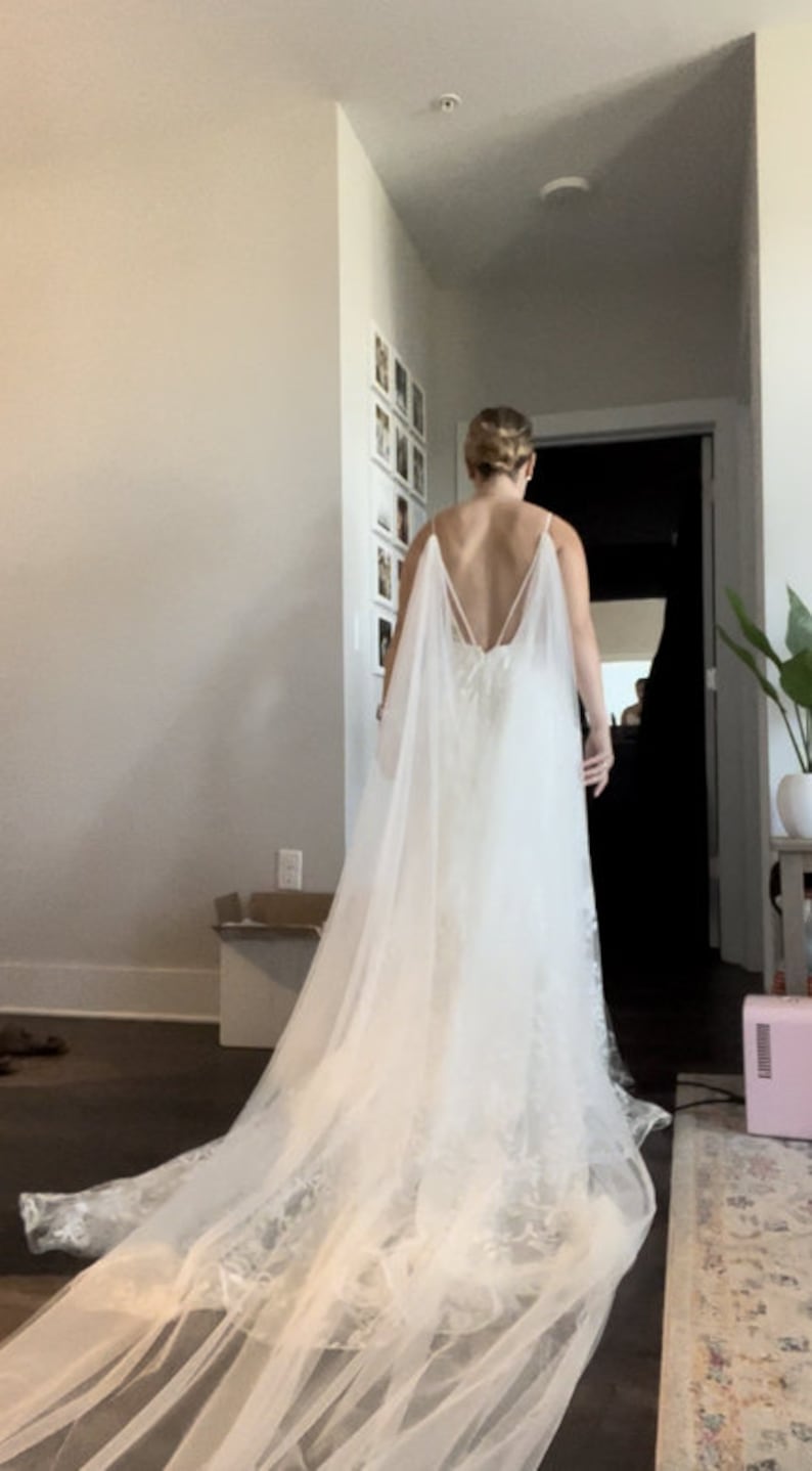 May include: A white wedding dress with a long train and a lace overlay. The dress has a low back and a cape-like overlay.