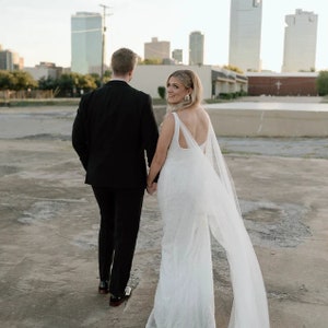 May include: A bride and groom walking away from the camera, the bride is wearing a white wedding dress with a long veil, the groom is wearing a black suit and black shoes. The couple is walking on a concrete surface with a city skyline in the background.