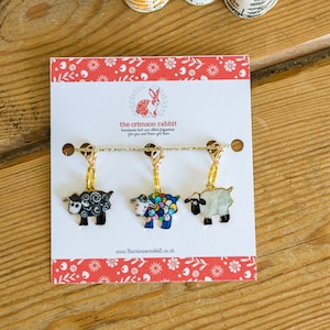 May include: Set of three sheep stitch markers with gold chains. The sheep are black and white, colourful, and white with black spots. The card reads "the crimson rabbit".
