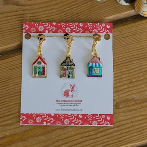May include: Set of three colourful house-shaped stitch markers with gold chains. The markers are on a white card with red polka dots and the text "the crimson rabbit handmade knit and stitch supplies for you and those you love www.thecrimsonrabbit.co.uk".