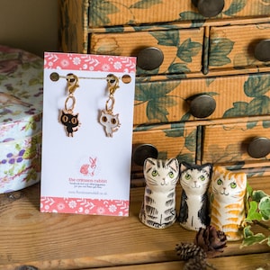 May include: Two gold cat stitch markers with black and white enamel details. The markers are hanging from a card with the text "the crimson rabbit" and a website address. Three ceramic cat figurines are in the background.