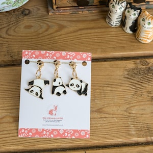 May include: Three gold panda bear charms with black and white enamel details. The charms are attached to gold lobster clasps. The charms are displayed on a white card with pink floral borders. The card has the text "the crimson rabbit" and a website address.