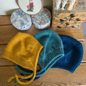 May include: Three knitted baby bonnets in mustard yellow, teal, and navy blue. The bonnets have a classic design with ties under the chin. The bonnets are displayed on a wooden surface, with decorative boxes and figurines in the background.