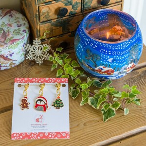 May include: A set of three gold-tone Christmas-themed stitch markers featuring a gingerbread person, a penguin wearing a red scarf, and a Christmas tree. The markers are on a white card with red and white stripes and the text "the crimson rabbit".