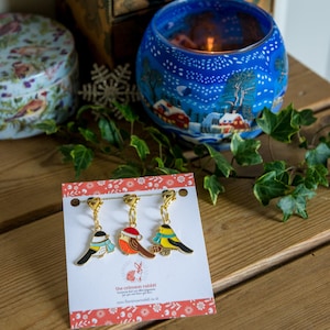May include: Three bird-shaped charms with gold-coloured clasps hang from a white card with a red border. The birds are painted in various colours, including blue, orange, and yellow, and one wears a Santa hat. The card has the text "the crimson rabbit" and a website address.