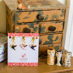 May include: Three gold bird charms with blue, pink, and green enamel. The charms are attached to a string and are hanging from a card with the text "The crimson rabbit" and a website address. The card is on a wooden surface with a small ceramic fox figurine and a vintage wooden drawer cabinet.