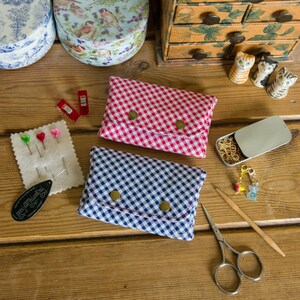 May include: Two small fabric pouches with gingham patterns. One pouch is red and white, the other is blue and white. Both pouches have a flap with a button closure. The pouches are on a wooden surface with other sewing supplies.