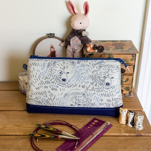 May include: A white zippered pouch with a blue trim and a pattern of white bears on a blue background. The pouch is sitting on a wooden table with knitting needles and a ruler.