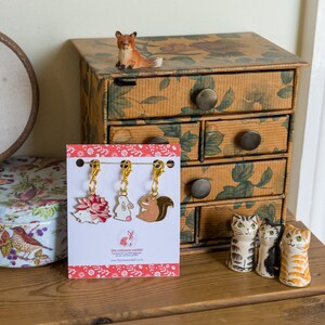 May include: Three gold-toned metal charms featuring a pink hedgehog, white rabbit, and brown squirrel design. The charms are attached to gold-toned clips and are displayed on a white card with a red and white floral pattern. The card includes the text "The Crimson Rabbit" and a website address.