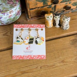 May include: Set of three gold-tone cat-themed stitch markers with enamel details. Each marker features a cat reading a book, with one cat wearing a hat and another holding a lamp. The markers are packaged on a white card with the text "the crimson rabbit" and a website address.
