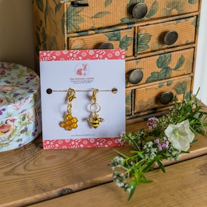 May include: A white card with a red and white floral border, featuring the text "the crimson rabbit" and a small illustration of a rabbit. The card is displaying two gold-coloured charms, one shaped like a honeycomb and the other like a bee. The charms are attached to gold-coloured clasps.