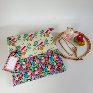 May include: A white and blue floral fabric pouch with a zipper closure. The pouch has a top section with a cream background and a repeating pattern of pink birds on branches. The pouch is sitting on a white surface with a wooden embroidery hoop, a ceramic cat figurine, and colourful embroidery floss.
