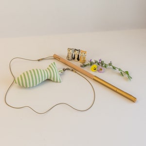 May include: A green and white striped fabric fish toy attached to a wooden and gold wand with a string. Three ceramic cat figurines are in the background.