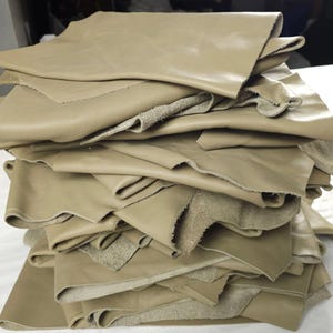May include: A stack of multiple irregular pieces of light tan leather. Each piece features a smooth, matte finish on the top side and a textured, suede-like surface on the underside. The edges of the leather pieces are raw, uneven, and unhemmed.