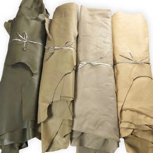 May include: Four rolls of leather in varying shades of earth tones, including olive green, tan, beige, and light cream. Each roll is secured with a thin silver metallic string tied in a simple bow, showcasing the natural texture and edges of the hide.