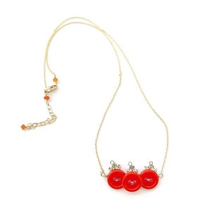 May include: A gold-colored necklace featuring three bright red button pendants. The buttons have a small, decorative design and are connected by a delicate gold chain. The necklace has an adjustable length and is set against a white background.