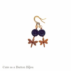May include: A pair of earrings featuring a dark blue button with a dragonfly charm hanging below. The earrings have a brass hook.