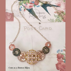May include: A gold-toned necklace featuring a series of decorative buttons in shades of pink, green, and gold. The central button is a large, openwork design encrusted with pink rhinestones. The text "Cute as a Button Bijou" is visible.