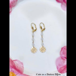May include: A pair of gold-colored dangle earrings. Each earring features a gold-colored lever back, a strand of small, light-colored beads, and a gold-colored circular charm with a floral design. The earrings are displayed on a white surface with pink floral accents. Text at the bottom reads "Cute as a Button Bijou."