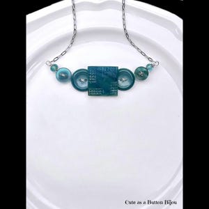 May include: A handmade necklace featuring a silver chain and teal-colored buttons. The necklace has a square button in the center, flanked by two round buttons, and smaller beads on each side. The text "Cute as a Button Bijou" is at the bottom.