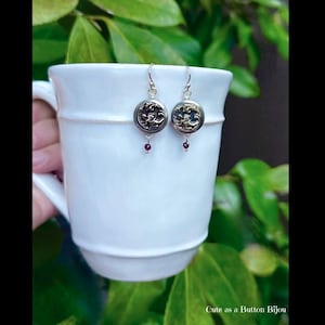 May include: A pair of button earrings with silver hooks. The earrings feature ornate, dark metal button faces with a floral design. Small, red beads dangle from the bottom of each earring. The earrings are displayed against a white mug.