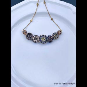 May include: A gold-toned necklace featuring a series of vintage buttons in various colors and designs. The buttons are arranged in a curved line, connected by a delicate chain. The necklace is displayed on a white surface. Text at the bottom reads "Cute as a Button Bijou."