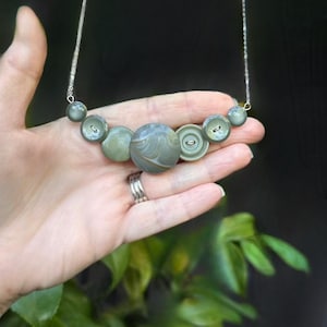 May include: A necklace featuring a series of seven sage green buttons of varying sizes arranged in a curved line. The buttons display swirling patterns and are connected by a thin silver-toned metal chain that extends upward from both ends.