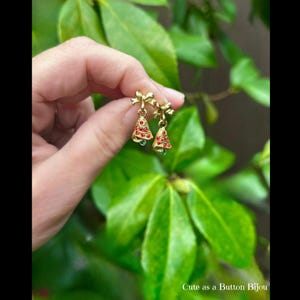 May include: Gold-toned Christmas tree earrings with red and green accents. Each earring features a small bow at the top and a bell-shaped tree design. The earrings are held against a backdrop of green leaves.