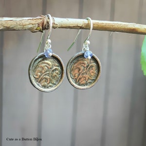 May include: A pair of silver dangle earrings with a round, antique-looking button charm featuring a floral design. Each button is accented with a small, light blue gemstone.