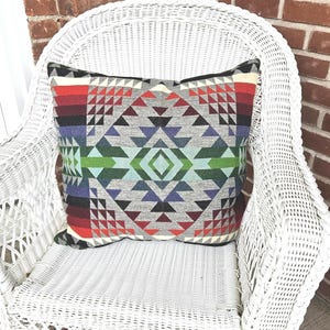 Op de afbeelding: Een vierkant sierkussen met een geometrisch patroon in grijstinten, rood, groen en paars ligt op een witte rotan stoel. Het ontwerp van het kussen bevat driehoeken en ruitvormen. De stoel staat op een patroonkleed.
