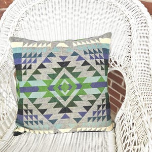 May include: A decorative square pillow with a geometric pattern in shades of gray, green, blue, and purple. The pillow is placed on a white wicker chair, creating a contrast of textures and colors. The pillow's design features triangles and diamond shapes.