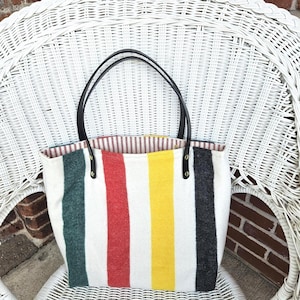 May include: A tote bag with a white background and vertical stripes in green, red, yellow, and black. The bag has black leather handles and a striped interior lining. The bag is sitting on a white wicker chair.