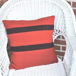 May include: A rectangular orange pillow with two wide, dark brown stripes. The pillow is placed on a white wicker chair, with a brick wall in the background. The pillow is a decorative item.