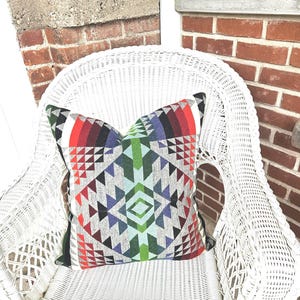 K&ouml;nnte beinhalten: Ein dekoratives Zierkissen mit geometrischem Muster in Rot, Gr&uuml;n, Blau und Grau liegt auf einem wei&szlig;en Korbstuhl. Das Kissen hat ein symmetrisches Design mit Dreiecken und Rauten, das dem neutralen Stuhl einen Farbtupfer verleiht.