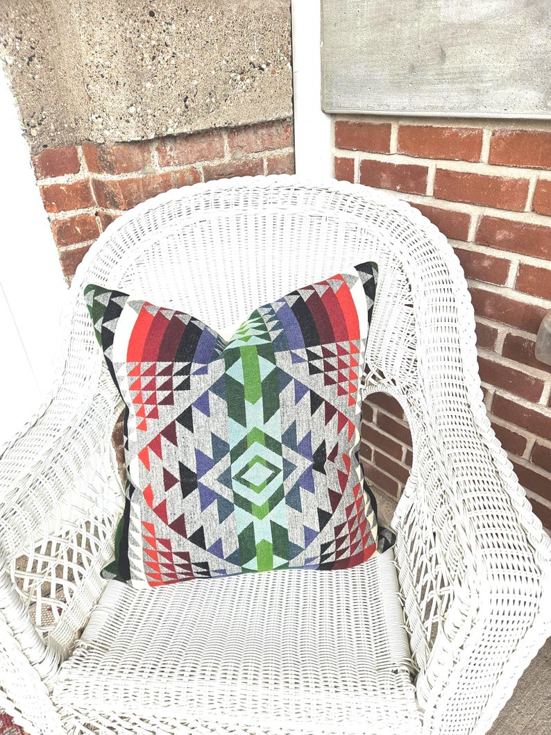 K&ouml;nnte beinhalten: Ein dekoratives Zierkissen mit geometrischem Muster in Rot, Gr&uuml;n, Blau und Grau liegt auf einem wei&szlig;en Korbstuhl. Das Kissen hat ein symmetrisches Design mit Dreiecken und Rauten, das dem neutralen Stuhl einen Farbtupfer verleiht.