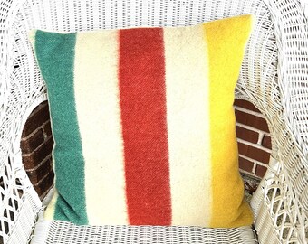 Puede incluir: Un cojín cuadrado con un patrón de rayas multicolor. El diseño consta de bandas verticales en tonos verde azulado, crema, rojo y amarillo. La tela tiene una apariencia suave y texturizada con un tejido visible en toda la superficie.