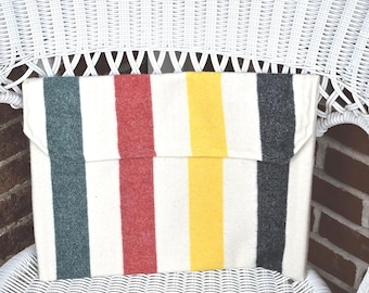 May include: A rectangular, off-white clutch with vertical stripes in green, red, yellow, and gray. The clutch is made of a soft, textured material and has a flap closure. It rests on a white wicker chair.