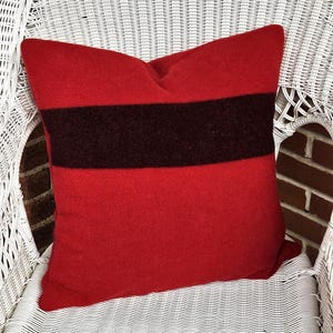 May include: A red pillow with a wide, dark brown stripe across the center. The pillow is made of a soft material and is placed on a white wicker chair. The pillow is square-shaped and appears to be a decorative item.