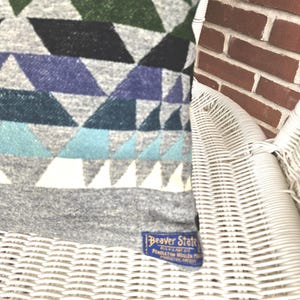 May include: A section of a patterned textile featuring geometric shapes in shades of gray, green, blue, and white. The textile has a label that reads "Beaver State". The textile is resting on a white wicker chair.