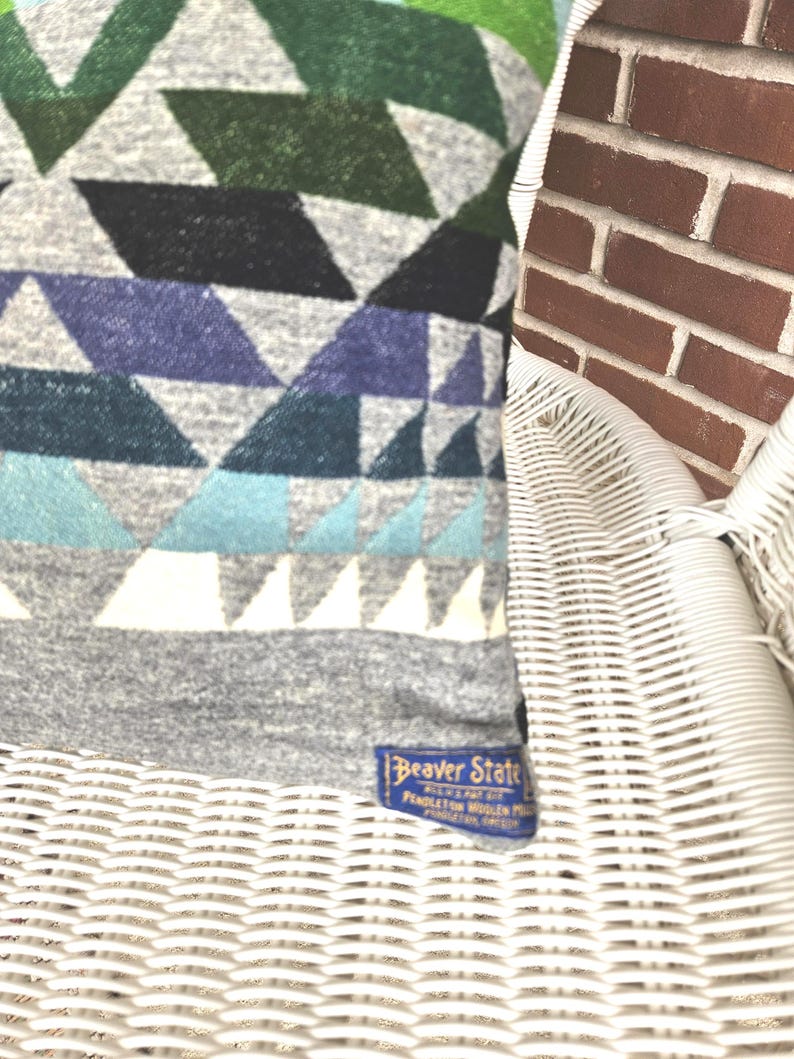 May include: A section of a patterned textile featuring geometric shapes in shades of gray, green, blue, and white. The textile has a label that reads "Beaver State". The textile is resting on a white wicker chair.