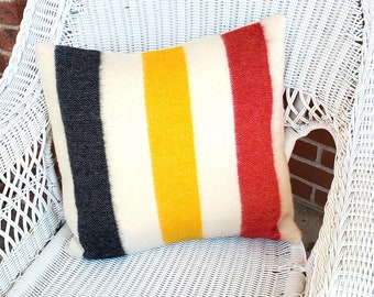 Puede incluir: Cojín cuadrado con un diseño de rayas en azul marino, amarillo y rojo. El cojín está hecho de una tela texturizada y descansa sobre una silla de mimbre blanca. Un elemento decorativo para el hogar.