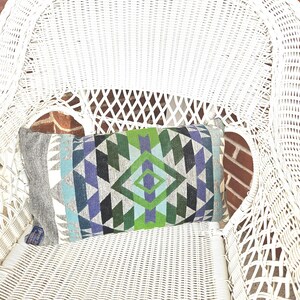 May include: A rectangular decorative pillow with a geometric pattern in shades of blue, green, gray, and black rests on a white wicker chair. The pillow's design features diamond and triangle shapes. The chair is positioned in front of a brick wall.