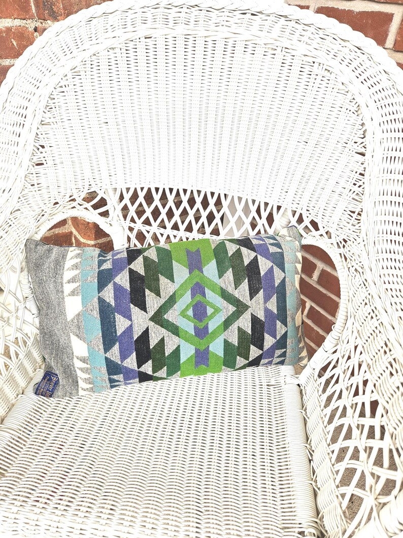 May include: A rectangular decorative pillow with a geometric pattern in shades of blue, green, gray, and black rests on a white wicker chair. The pillow's design features diamond and triangle shapes. The chair is positioned in front of a brick wall.