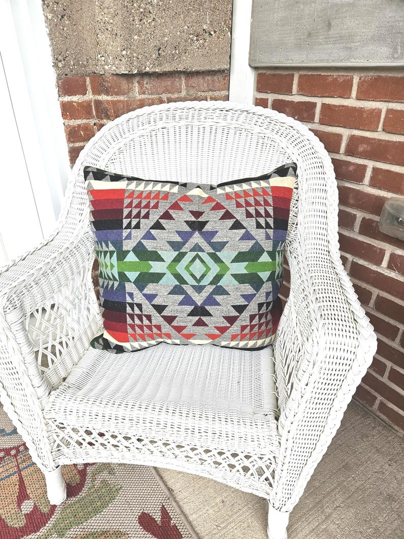 K&ouml;nnte beinhalten: Ein quadratisches Zierkissen mit einem geometrischen Muster in Grau-, Rot-, Gr&uuml;n- und Lilat&ouml;nen liegt auf einem wei&szlig;en Korbstuhl. Das Design des Kissens zeigt Dreiecke und Rauten. Der Stuhl steht auf einem gemusterten Teppich.