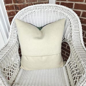 K&ouml;nnte beinhalten: Ein cremefarbenes Kissen liegt auf einem wei&szlig;en Korbstuhl. Das Kissen hat ein weiches, strukturiertes Aussehen und ein gefaltetes Design. Das komplizierte Webmuster des Stuhls und der Ziegelhintergrund tragen zur gem&uuml;tlichen &Auml;sthetik des Bildes bei.