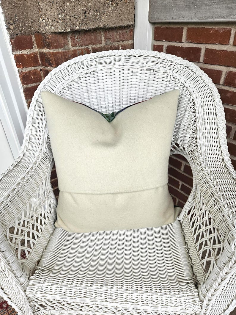 K&ouml;nnte beinhalten: Ein cremefarbenes Kissen liegt auf einem wei&szlig;en Korbstuhl. Das Kissen hat ein weiches, strukturiertes Aussehen und ein gefaltetes Design. Das komplizierte Webmuster des Stuhls und der Ziegelhintergrund tragen zur gem&uuml;tlichen &Auml;sthetik des Bildes bei.