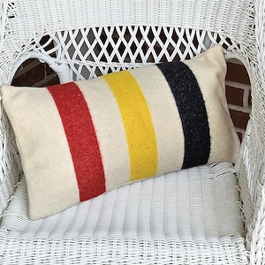 May include: A rectangular pillow with a cream-colored background and bold vertical stripes in red, yellow, and black. The pillow is placed on a white wicker chair, creating a cozy and inviting aesthetic.