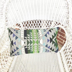 May include: Rectangular decorative pillow with a geometric pattern in shades of gray, green, blue, and white. The pillow is resting on a white wicker chair, creating a contrast of textures and colors. The pillow's design features triangles and other shapes.